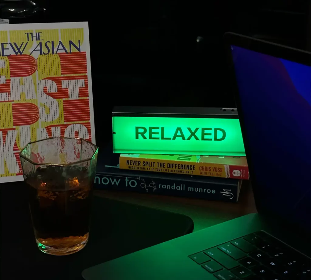 A green mood light with 'RELAXED' sign, a dimly lit desk with a stack of books, a glass of amber liquid, and a laptop keyboard.