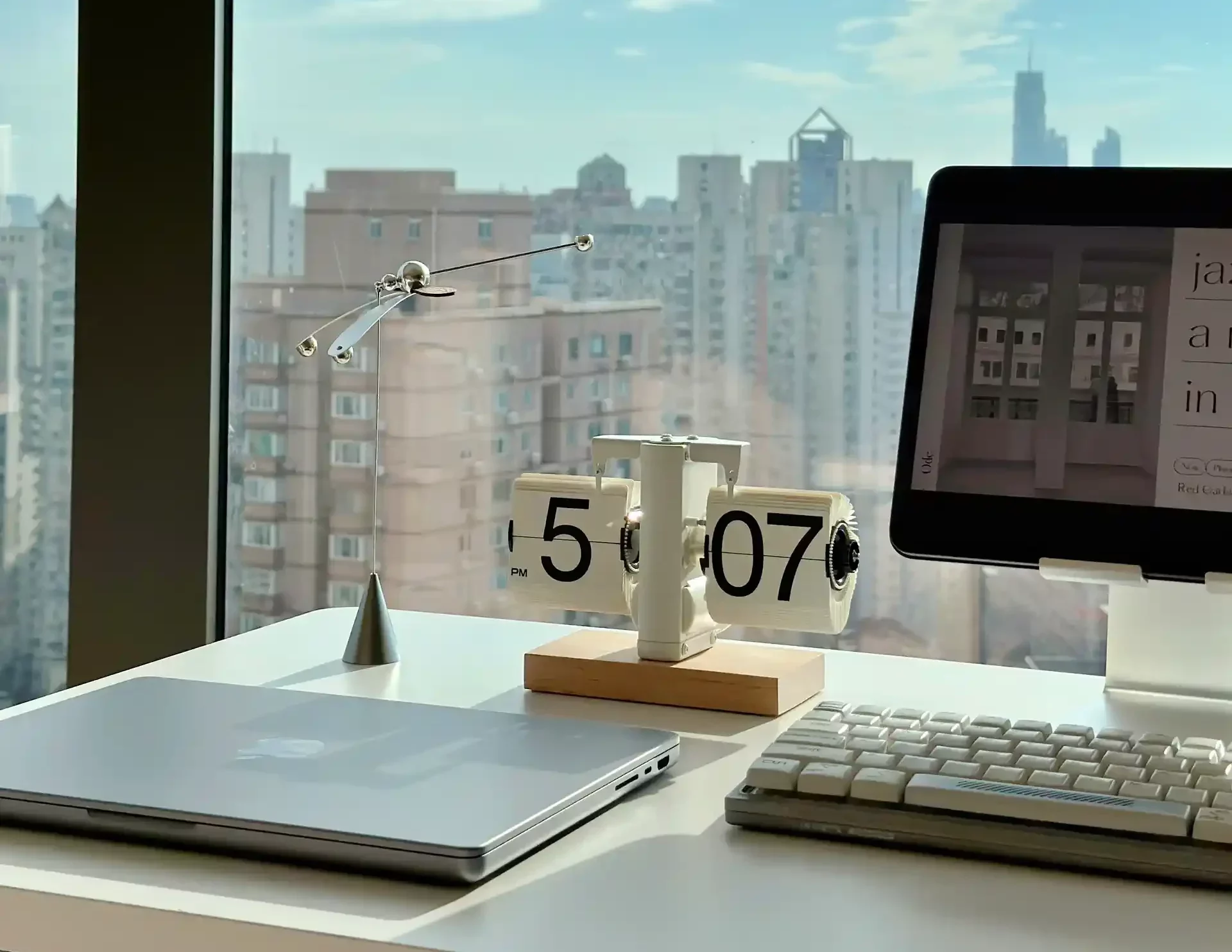 A modern workspace beside a window, with a laptop, digital clock displaying 5:07 PM, and retro style keyboard, and a metallic articulating lamp.
