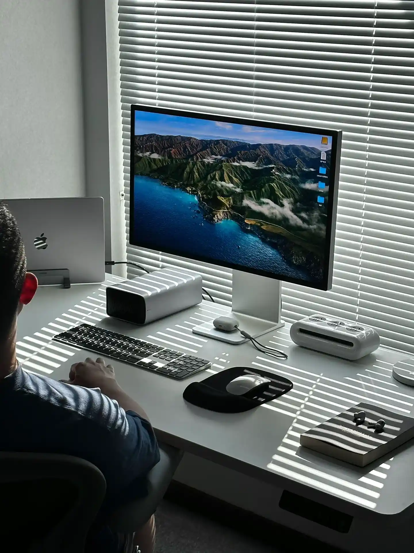 Modern desk decor setup for guys with a monitor displaying scenic mountains, Apple devices, striped blinds casting shadows, and minimalist accessories