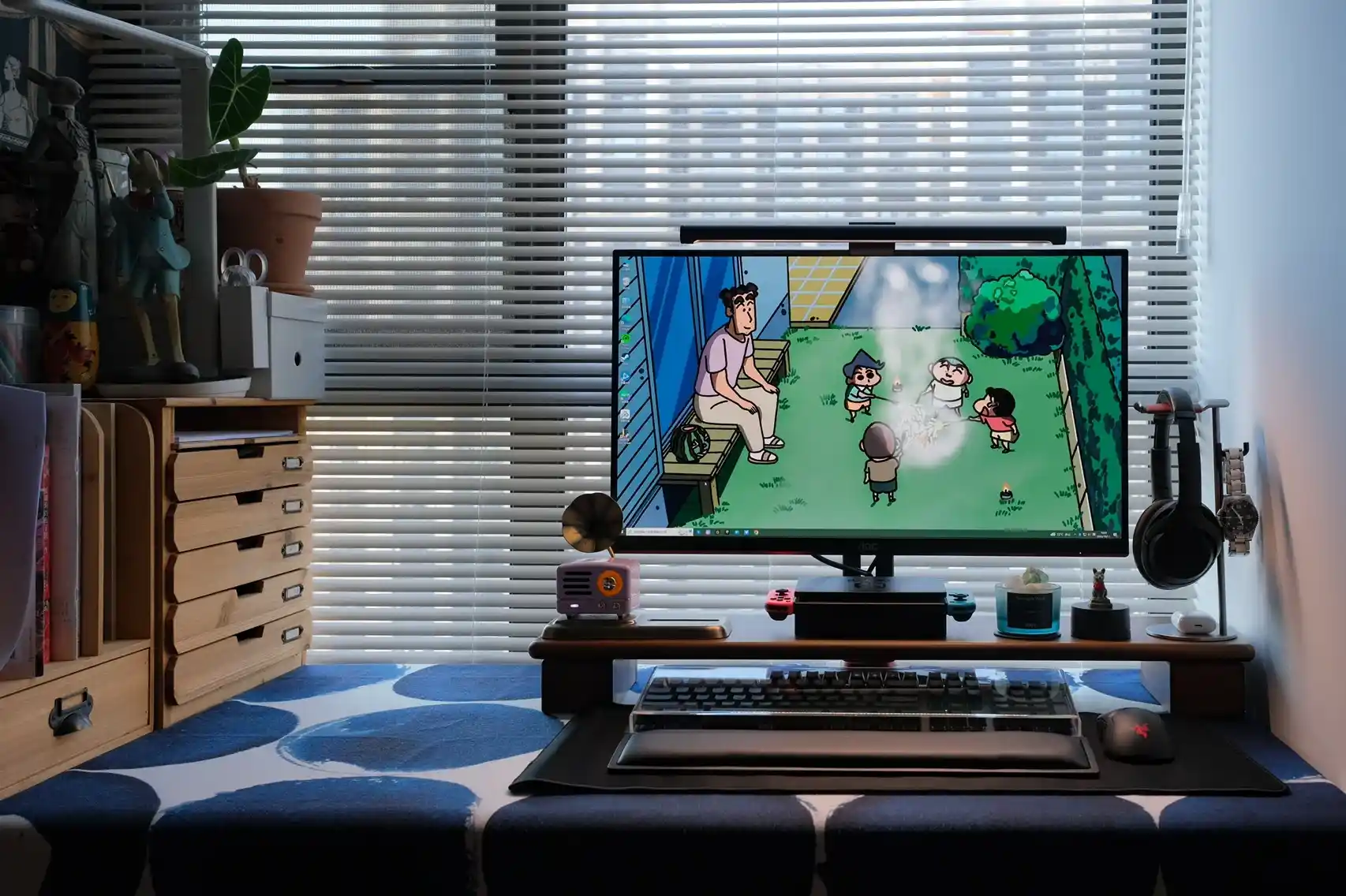 A cozy workspace featuring a computer monitor displaying animated characters, surrounded by various desk accessories, plants, and figurines.