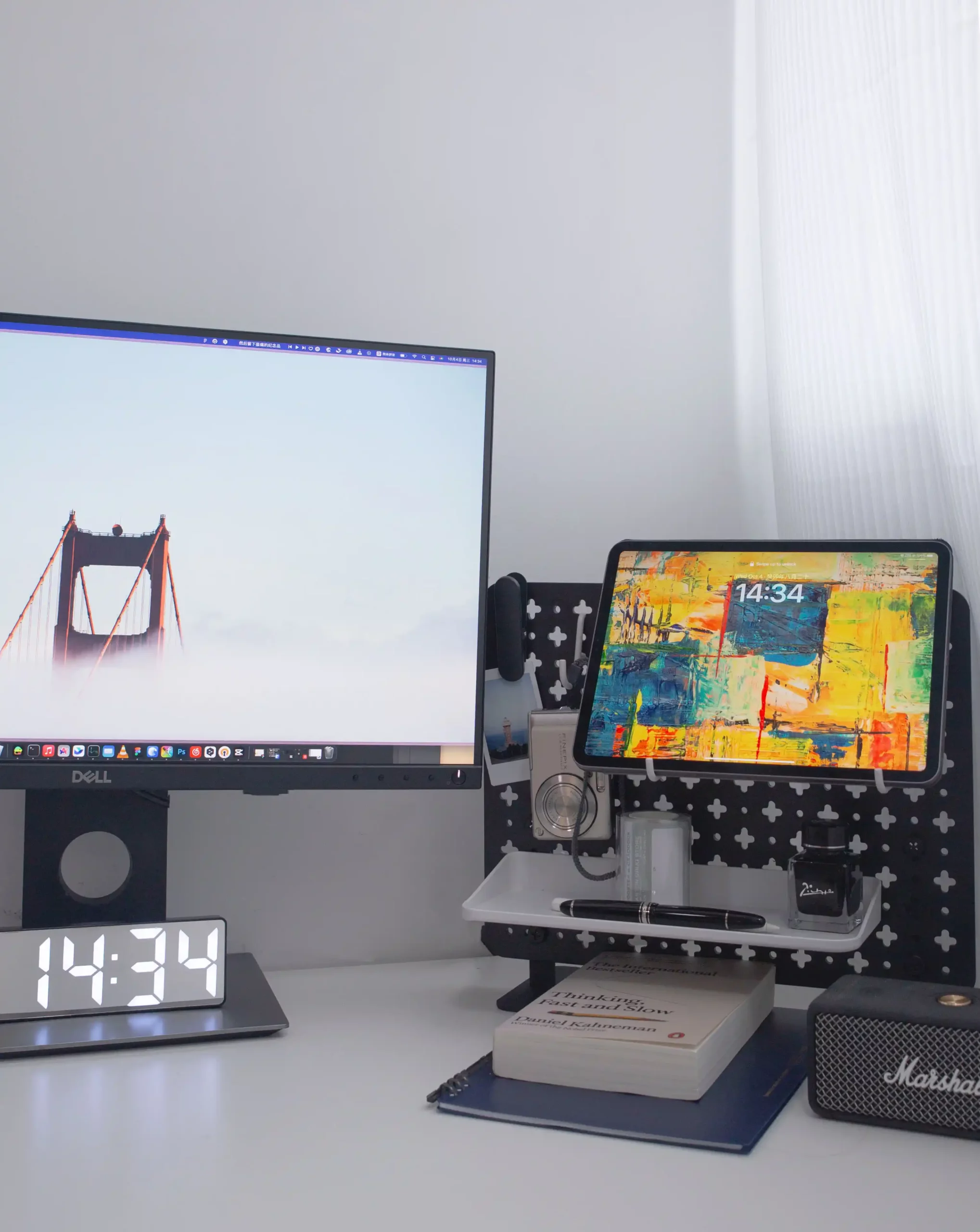A minimalistic workstation with a computer monitor displaying an image of a bridge, an iPad with an abstract art wallpaper, and assorted desk accessories including a digital clock displaying 14:34.