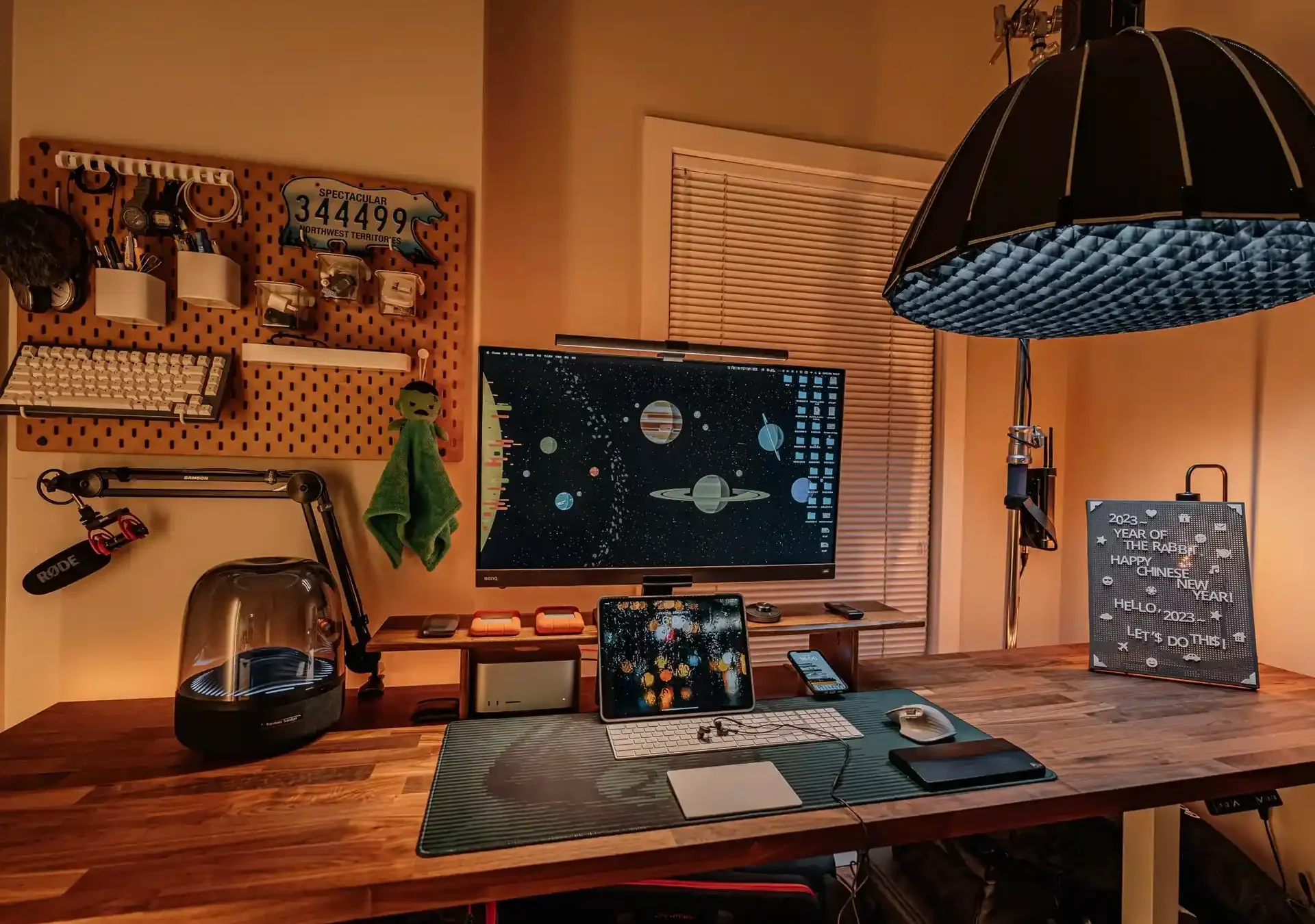 A cozy home office setup featuring a wooden desk with a computer displaying a space-themed wallpaper, various gadgets, and a pegboard with assorted items.